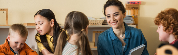heureux jeune enseignant souriant près de interracial enfants dans montessori école, bannière - Photo, image