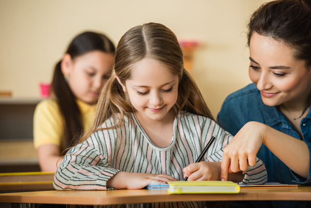 profesor apuntando cerca sonriente niño escritura en notebook y asiático chica en borrosa fondo - Foto, Imagen