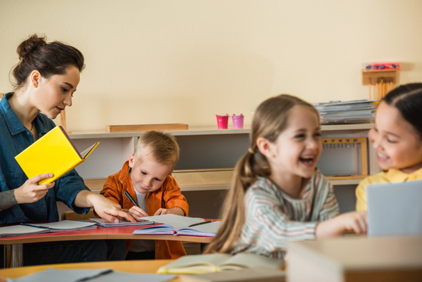 teacher pointing at notebook near writing boy while interracial girls laughing on blurred foreground - Foto, afbeelding