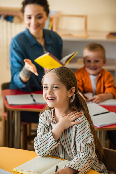blurred teacher pointing at girl smiling in classroom in montessori school  - Foto, afbeelding