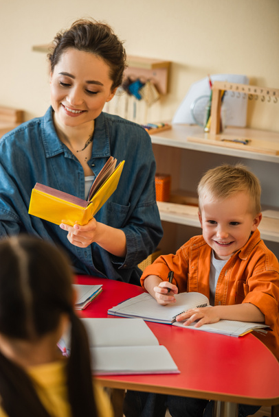 Улыбающийся учитель держит книгу рядом с счастливым мальчиком и размытой девушкой в школе Монтессори - Фото, изображение