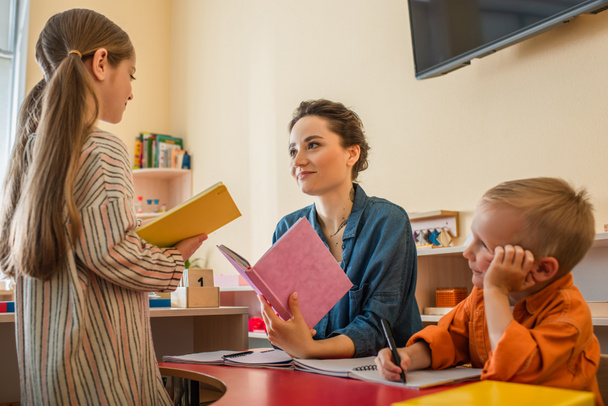 lány könyv áll közel a tanár és a fiú ül az asztalnál az osztályteremben - Fotó, kép