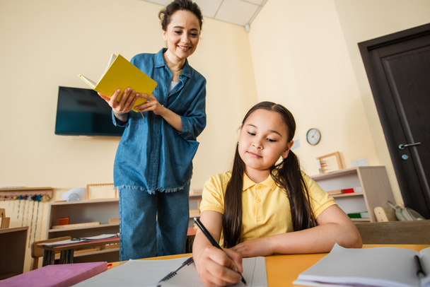 щасливий вчитель дивиться на азіатську дівчину, що пише в блокноті під час уроку в школі Монтессорі
 - Фото, зображення