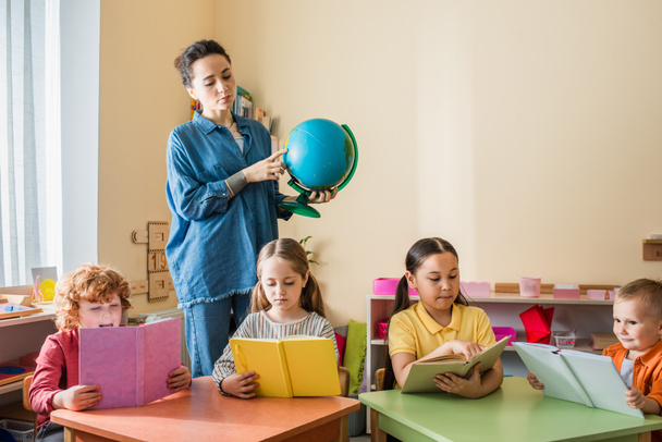 Lehrerin zeigt auf Globus in der Nähe multiethnischer Kinder, die während des Unterrichts Bücher lesen - Foto, Bild