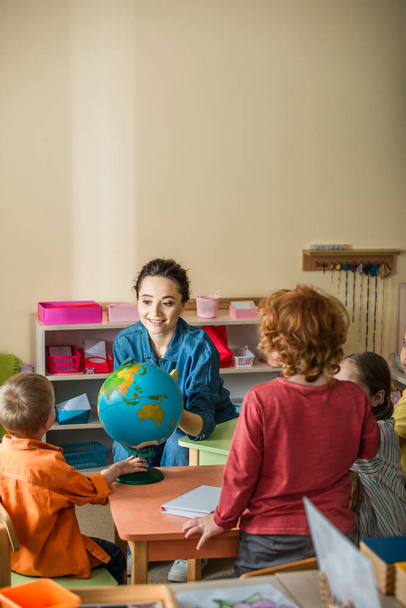 усміхнений вчитель розмовляє з дітьми поблизу земної кулі в класі
 - Фото, зображення