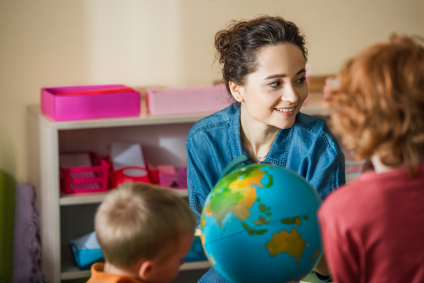 terug zicht op wazige jongens in de buurt globe en glimlachende leraar in Montessori school - Foto, afbeelding