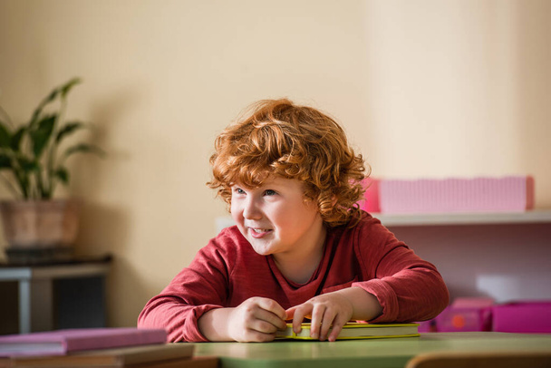 happy, curly boy smiling near blurred books in montessori school - Photo, Image
