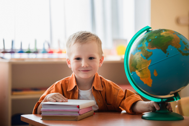 cheerful boy looking at camera near books and globe on desk - Fotó, kép