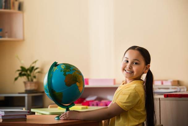 joyful asian girl smiling at camera near globe in montessori school - Photo, Image
