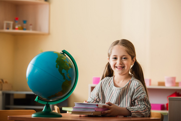 happy kid sitting with books near globe in montessori school - Photo, Image