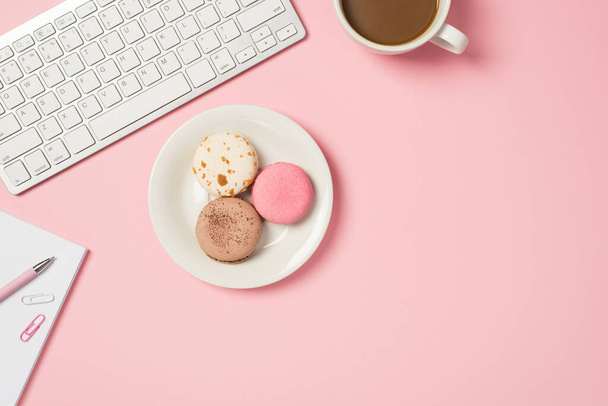 Над фото клавиатуры чашки кофе ноутбук с ручкой скрепки бумаги и пластины различных макарон изолированы на розовом фоне с копирайтом - Фото, изображение