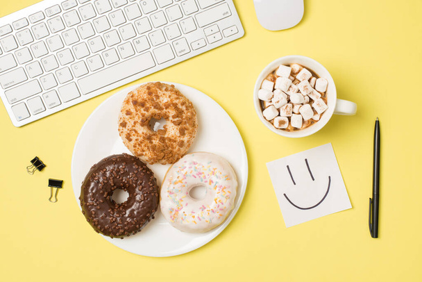 Top view photo of workplace white keyboard mouse binder clips pen sticker note with drawn smiling face cup of drink with marshmallow and plate with three donuts on isolated light yellow background - Photo, Image