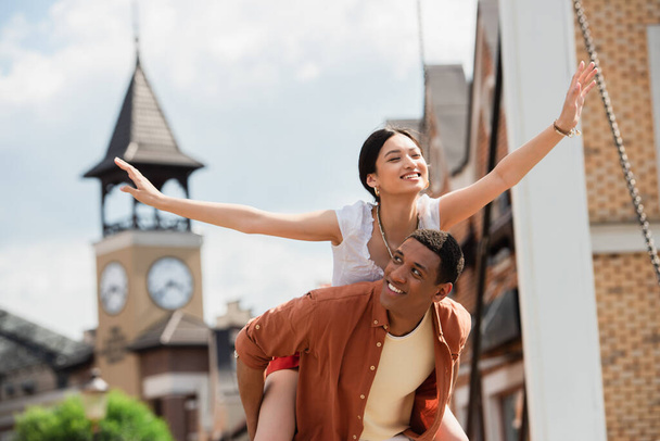 cheerful asian woman piggybacking on happy african american man outdoors - Foto, immagini