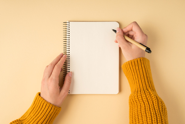Primeira pessoa vista superior foto de mãos femininas em pulôver amarelo escrevendo em bloco de notas em espiral em fundo laranja claro isolado com copyspace - Foto, Imagem