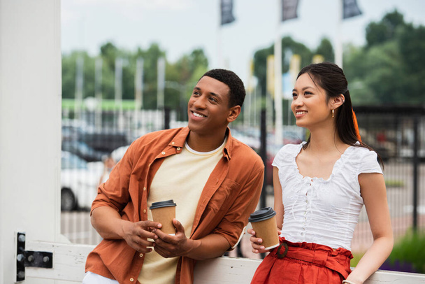 stylish and happy multiethnic couple holding coffee to go while looking away outdoors - Foto, Imagen