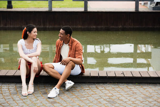 happy  interracial couple talking while sitting on border near water - Photo, Image