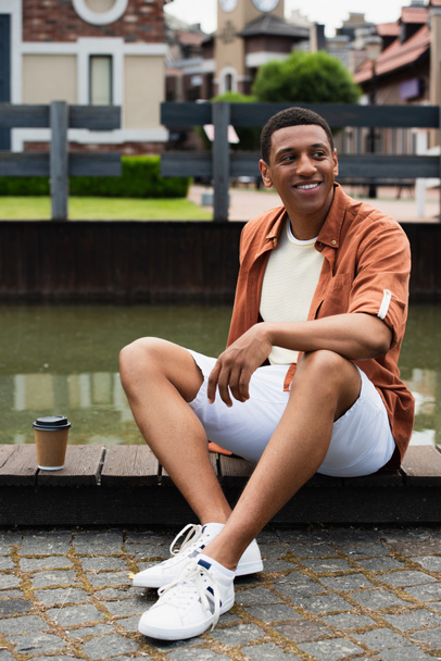 happy african american man sitting on border near water and coffee to go on urban street - Photo, image