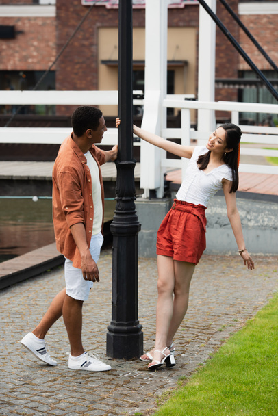 elegante casal interracial olhando uns para os outros perto lanterna na cidade - Foto, Imagem