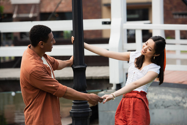 happy interracial couple holding hands and looking at each other near lantern - Photo, image