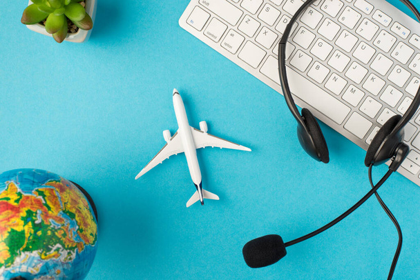Horní pohled fotografie černé sluchátka s mikrofonem na bílé klávesnici květináč globus a letadlo model uprostřed na izolované pastelové modré pozadí - Fotografie, Obrázek