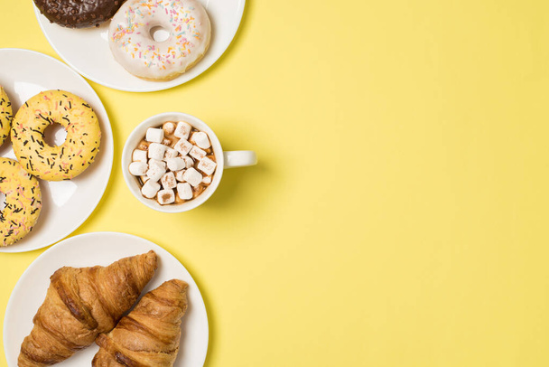 Top view photo of cup of drink with marshmallow and three plates with different color glazed donuts and fresh croissants on isolated pastel yellow background with empty space - Photo, Image
