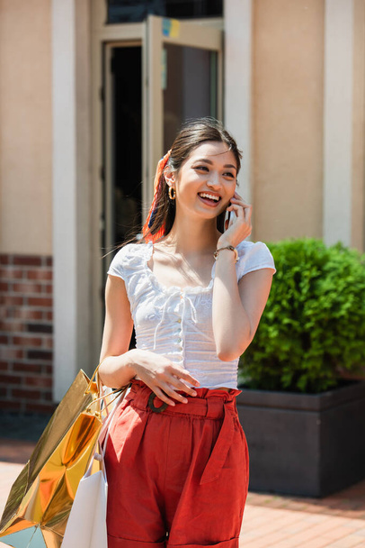 joyful asian woman with shopping bags talking on mobile phone outdoors - Fotoğraf, Görsel
