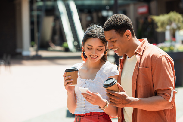 cheerful interracial couple with paper cups looking at mobile phone outdoors - Foto, imagen