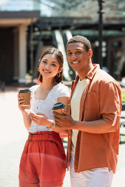 cheerful interracial couple with coffee to go smiling at camera outdoors - Fotó, kép