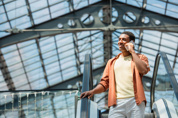 mosolygós afro-amerikai férfi néz félre, miközben beszél okostelefonon mozgólépcsőn - Fotó, kép