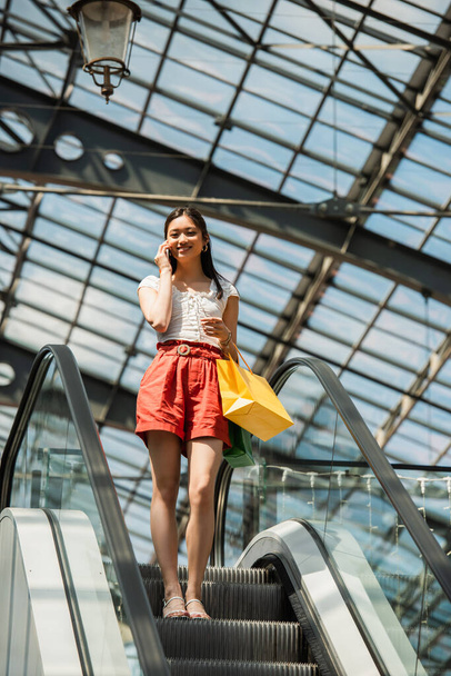 vidám ázsiai nő vásárlások beszél okostelefon mozgólépcsőn - Fotó, kép