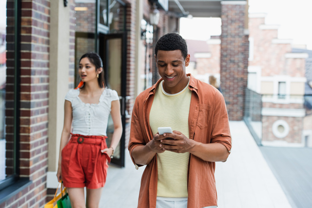 fröhliche afrikanisch-amerikanische Mann mit Handy in der Nähe asiatische Freundin mit Einkaufstüten - Foto, Bild