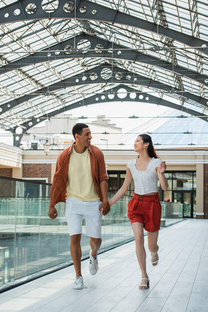 excited multiethnic couple holding hands while running under glass roof in city - Фото, изображение