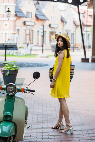 bonita mujer asiática en amarillo vestido de sol y sombrero mirando la cámara cerca de scooter - Foto, Imagen