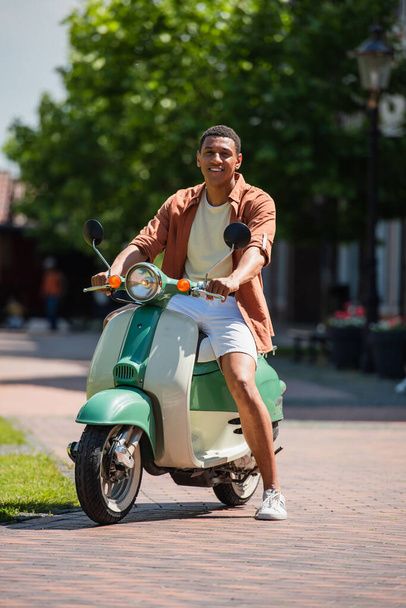 młody Afrykański Amerykanin na skuterze uśmiecha się do kamery na miejskiej ulicy - Zdjęcie, obraz