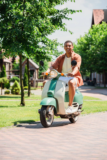pozytywny Afryki amerykański człowiek patrząc w aparat podczas jazdy skuter na zielonej ulicy - Zdjęcie, obraz