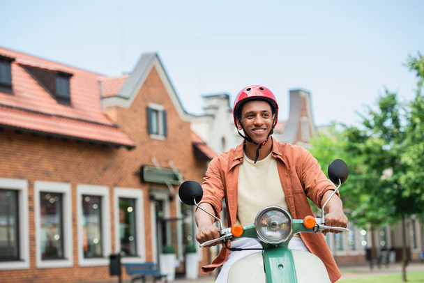 sonriente afroamericano hombre en casco a caballo scooter en la ciudad - Foto, Imagen