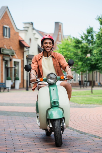 hymyilevä afrikkalainen amerikkalainen mies kypärässä nauttien ratsastus skootteri kaupungissa - Valokuva, kuva