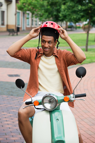positive african american man putting on hardhat while sitting on scooter - Φωτογραφία, εικόνα