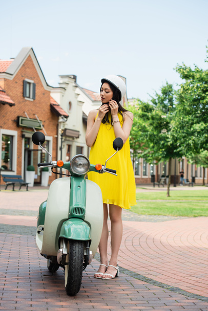 young asian woman in yellow sundress fastening helmet near scooter outdoors - Photo, Image