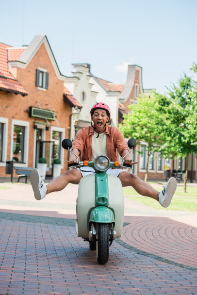 thrilled african american man raising legs while riding scooter outdoors - Fotoğraf, Görsel