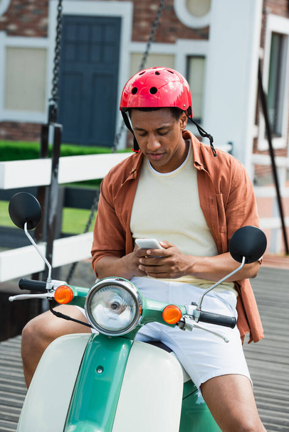 Αφρικανός Αμερικανός άνθρωπος σε κράνος μηνυμάτων σε smartphone, ενώ κάθεται σε σκούτερ - Φωτογραφία, εικόνα