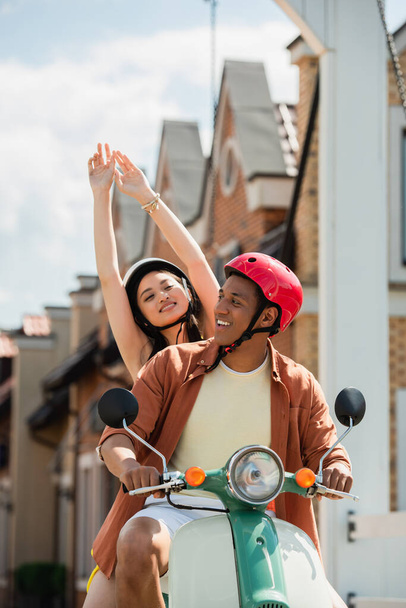 feliz mulher asiática com as mãos levantadas desfrutando equitação scooter com Africano americano homem - Foto, Imagem