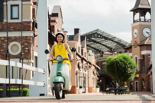 joven asiático mujer en amarillo sundress y casco a caballo scooter en ciudad calle - Foto, imagen