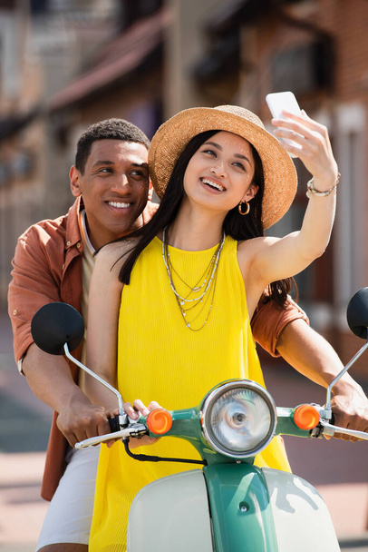 joyful asian woman taking selfie with african american boyfriend on scooter - Zdjęcie, obraz