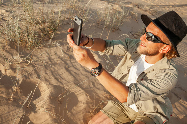un hombre con gafas y un sombrero en el desierto en el verano toma fotos por teléfono - Foto, Imagen