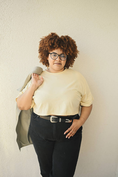 Beautiful happy smiling curvy plus size African black woman afro hair posing beige t-shirt, jeans, stylish glasses on beige background. Body imperfection, acceptance body positive diversity concept - Photo, Image