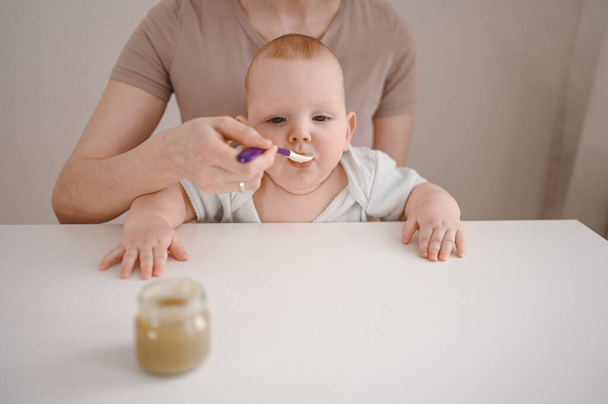 Little newborn funny baby boy learning to eat vegetable or fruit puree from glass jar with spoon. Young mother helping little son eat first food. - Фото, изображение