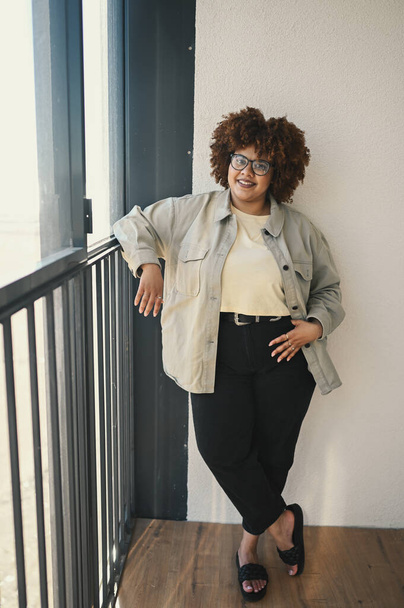 Beautiful happy smiling curvy plus size African black woman afro hair posing in beige t-shirt, jeans, stylish glasses on sunny balcony. Body imperfection, acceptance body positive diversity concept - Foto, Imagem