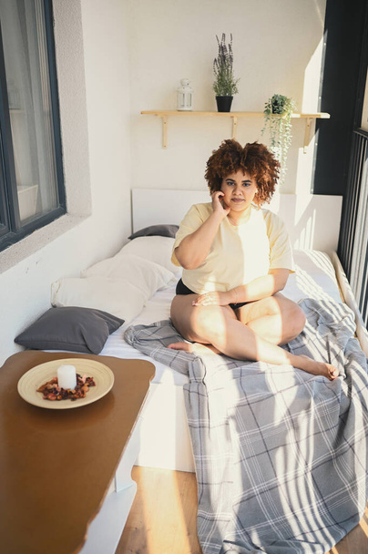 Piękny szczęśliwy curvy plus size Afrykański czarny kobieta afro włosy pozowanie w beżowym t-shirt i bielizna na słoneczny balkon sypialnia. Niedoskonałość ciała, akceptacja ciała, koncepcja ciała pozytywnego i różnorodności - Zdjęcie, obraz