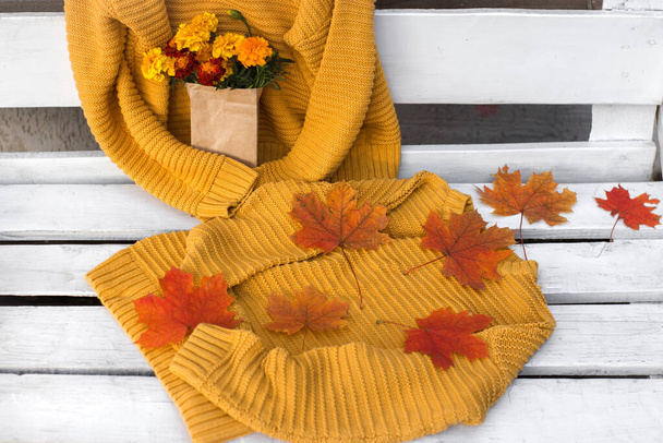 egy fehér fa padon, két egyforma kötött világos sárga pulóver, őszi juharfalevél, egy csokor kerti virág egy kézműves táskában. Őszi hangulat - Fotó, kép